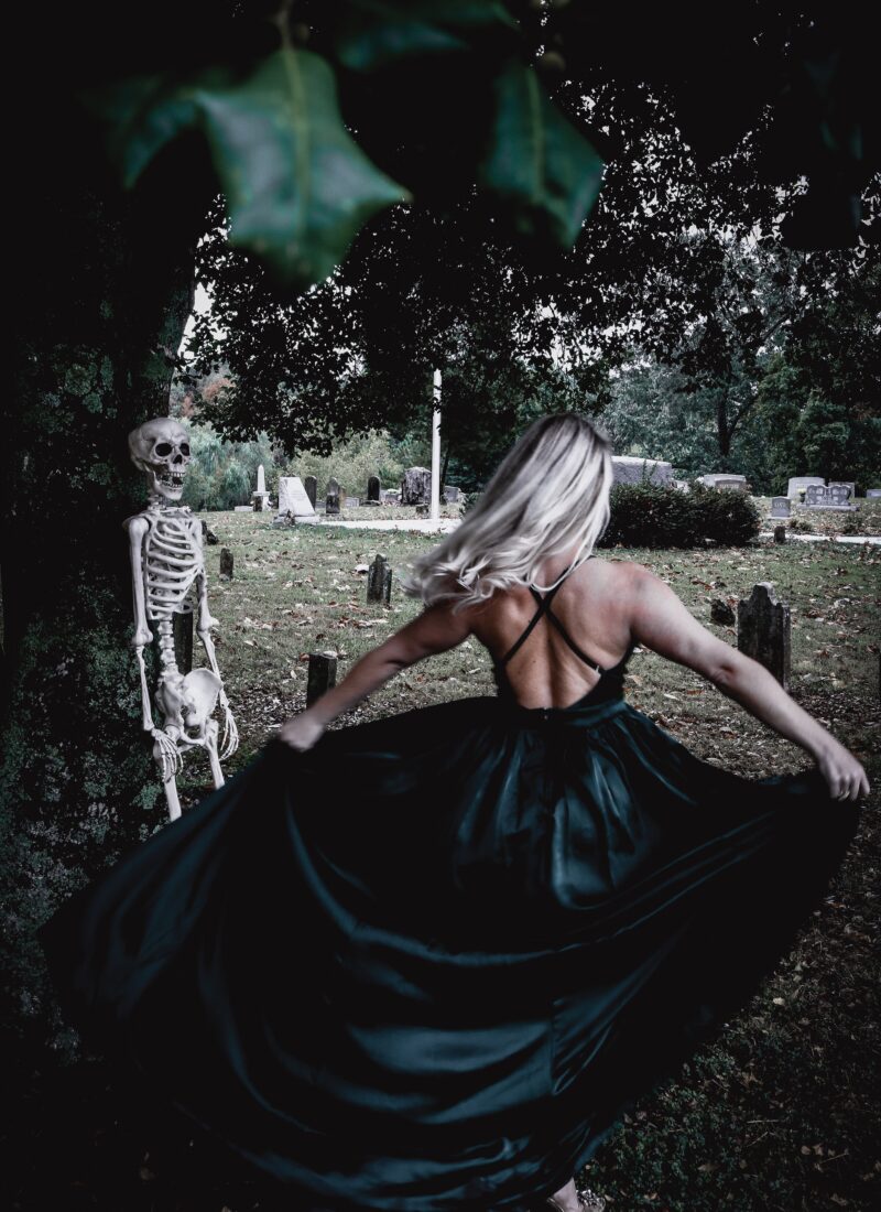 Photograph of Stephanie Carroll twirling in a graveyard with a skeleton during spooky season.